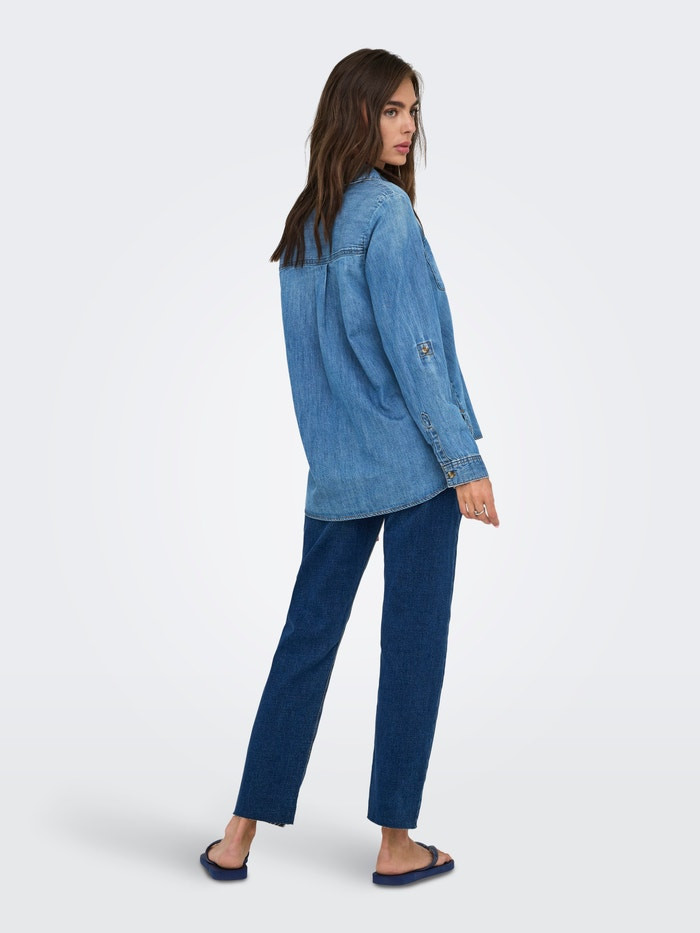 Side back view of model in ONLYASMIN blue denim shirt and dark jeans, walking on white floor