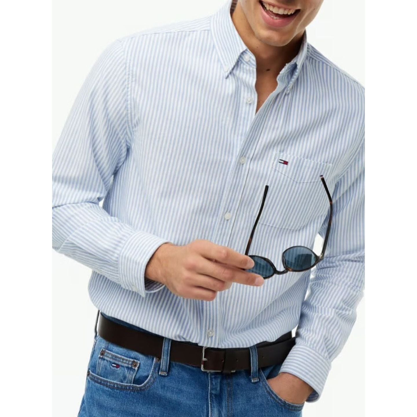 Gros plan sur la chemise Oxford à rayures portée ouverte au col, le mannequin tenant des lunettes pr