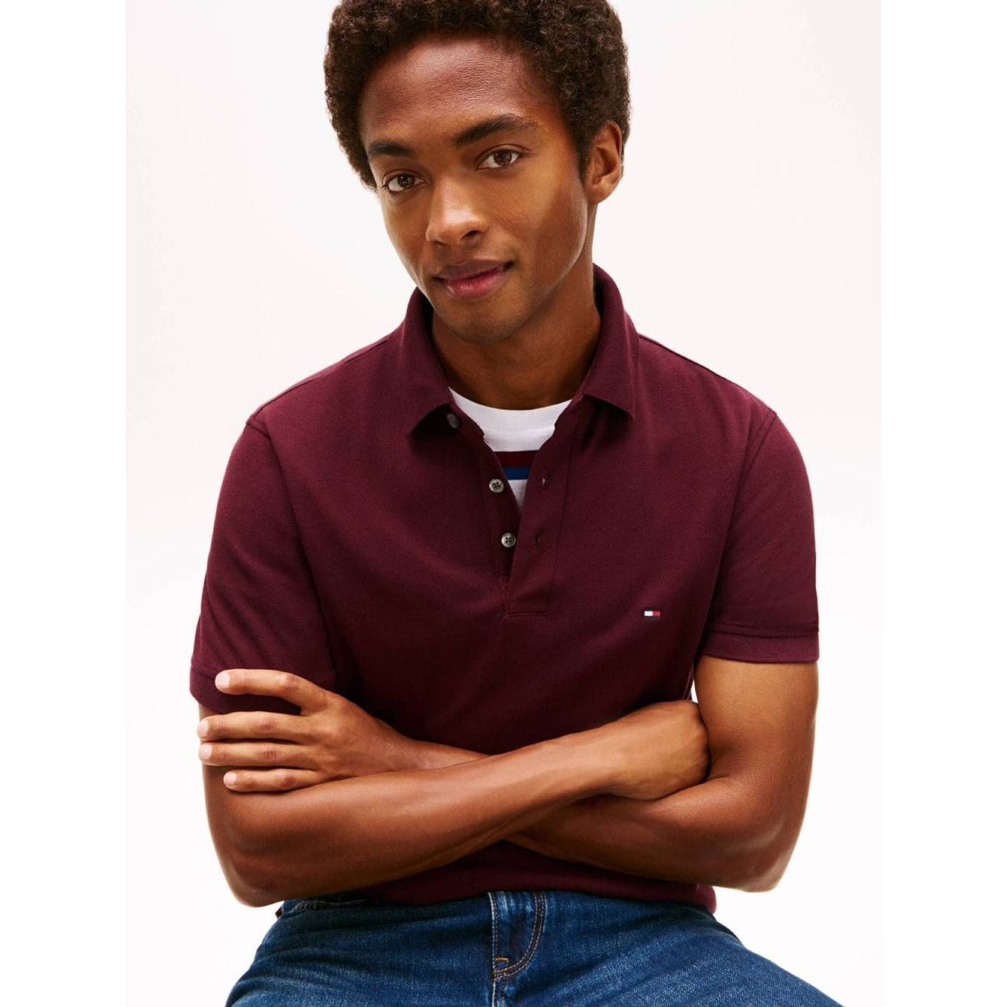 Homme assis, bras croisés, portant le polo Tommy Jeans Purple Berry superposé à un tee-shirt rayé bl
