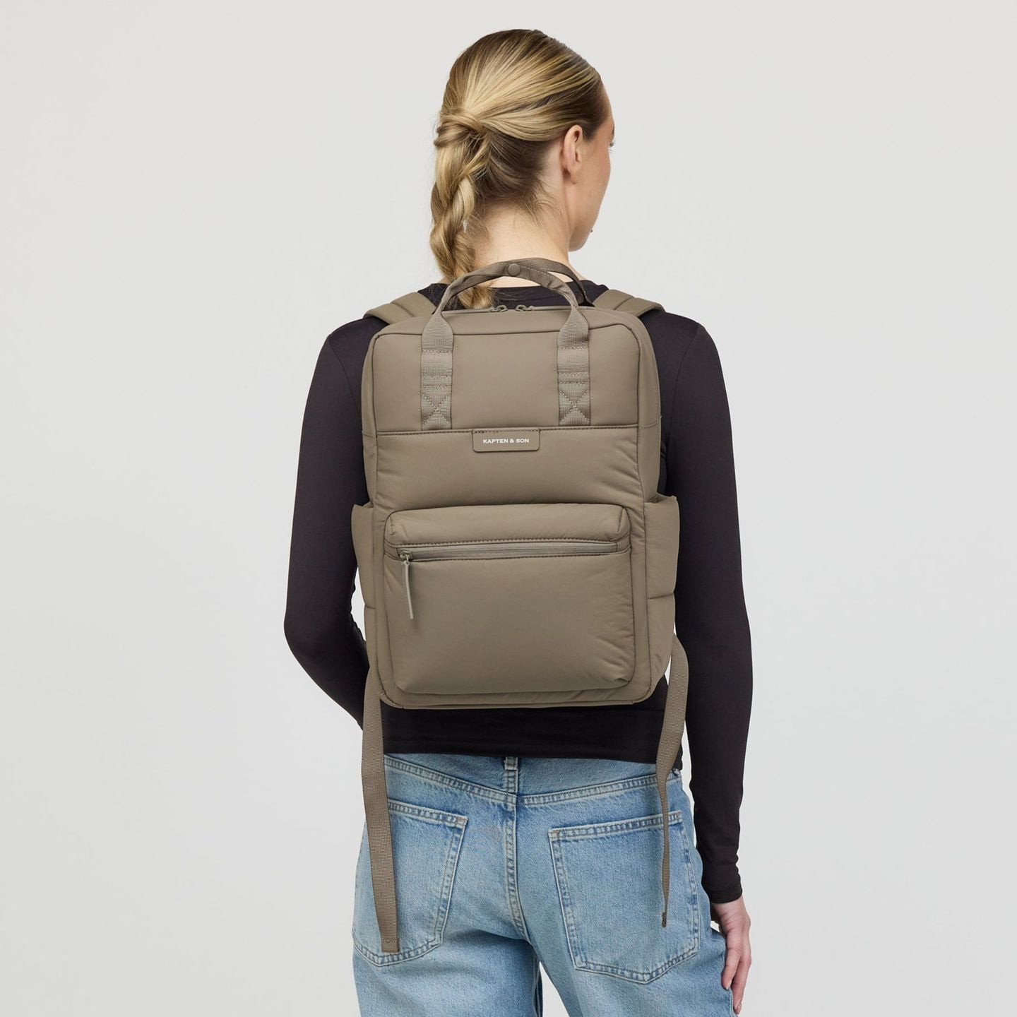 Femme portant le sac à dos Bergen Cloud Walnut sur un haut noir et un jean, vue de dos sur fond clai