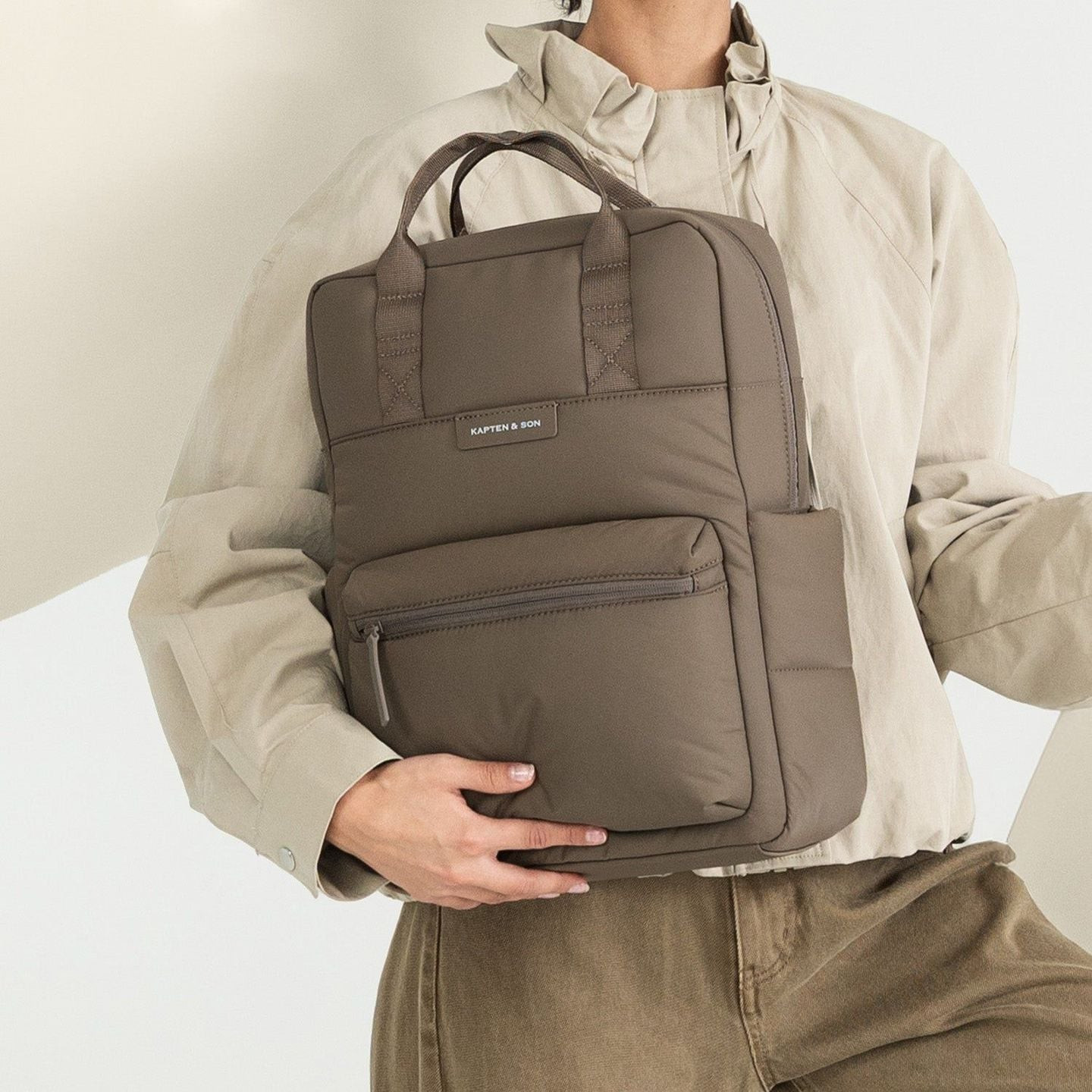 Modèle tenant le sac Bergen Cloud Walnut par la poignée supérieure contre une veste beige, taille co