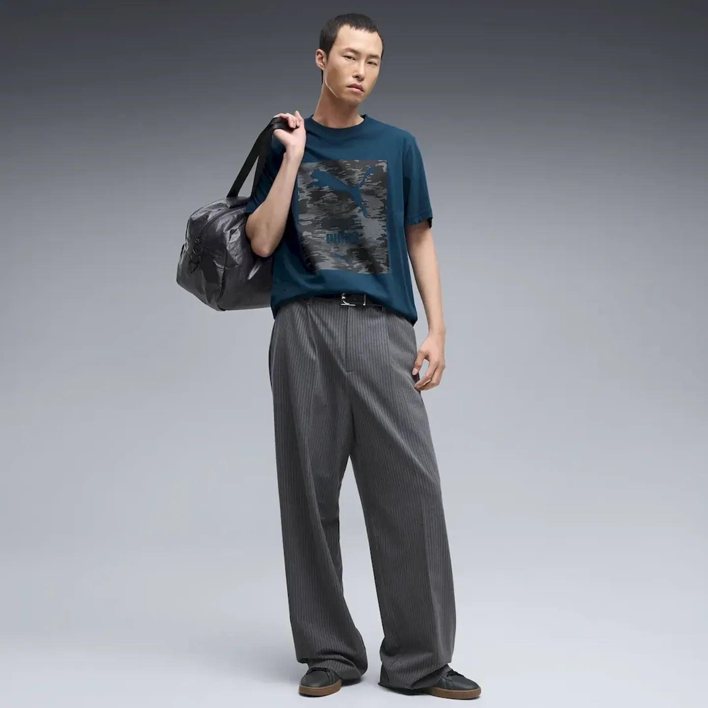 Vue en pied du mannequin portant un t-shirt PUMA Sport Camo Graphic Tee, pantalon gris et sac à l'ép