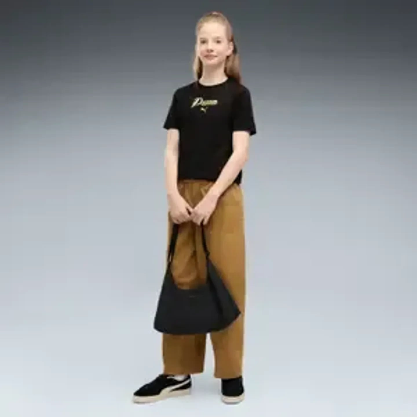 Fille en tee-shirt PUMA ESS Script Glitter G noir, logo doré, pantalon marron, sac noir et baskets.