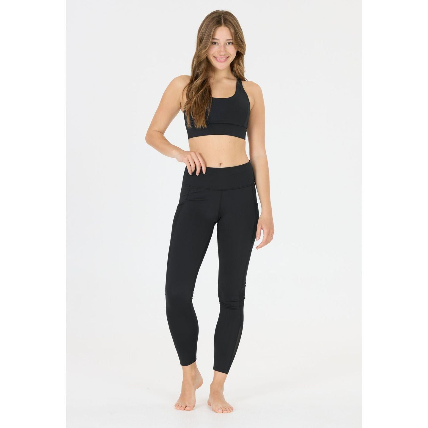 Mannequin souriant en brassière de sport Athlecia DIRENNA noire et legging noir, pieds nus sur fond 