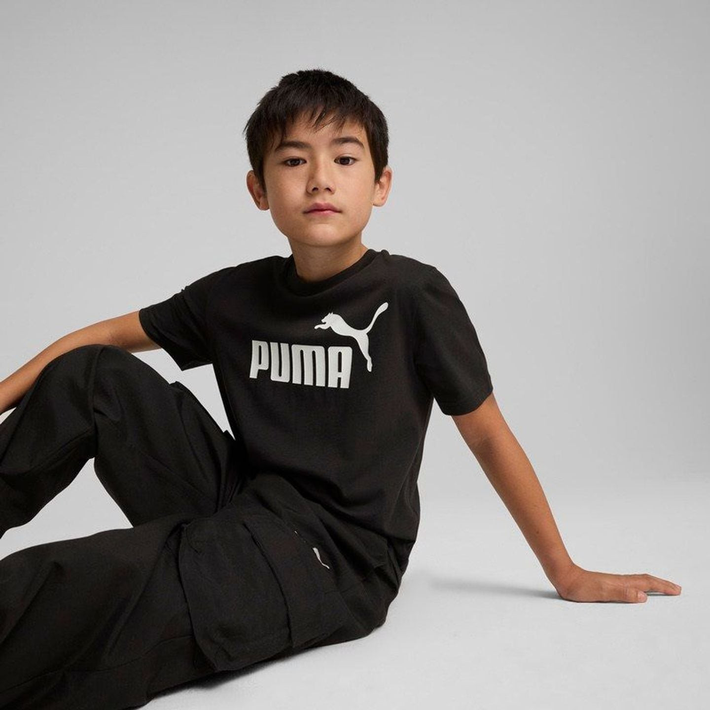 Garçon junior assis en studio portant le tee-shirt PUMA ESS No. 1 Logo Tee B noir avec logo blanc su