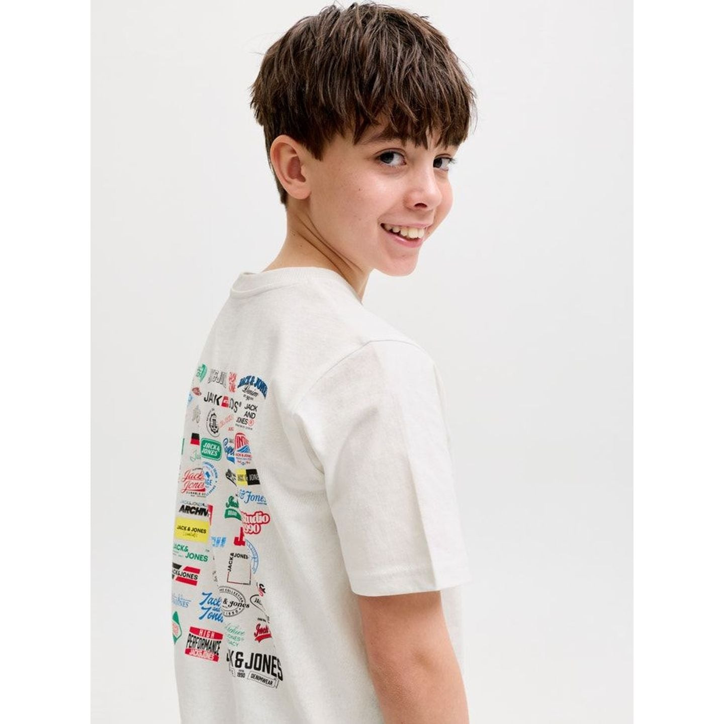 Garçon portant le t-shirt blanc JACK & JONES JUNIOR, souriant par-dessus son épaule pour montrer l'i