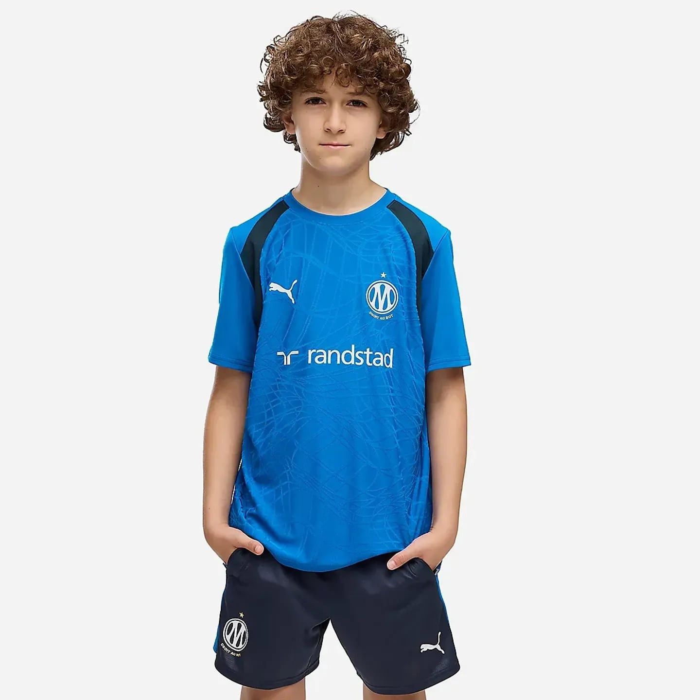 Jeune garçon portant le maillot d'entraînement junior OM bleu et un short marine, mains dans les poc