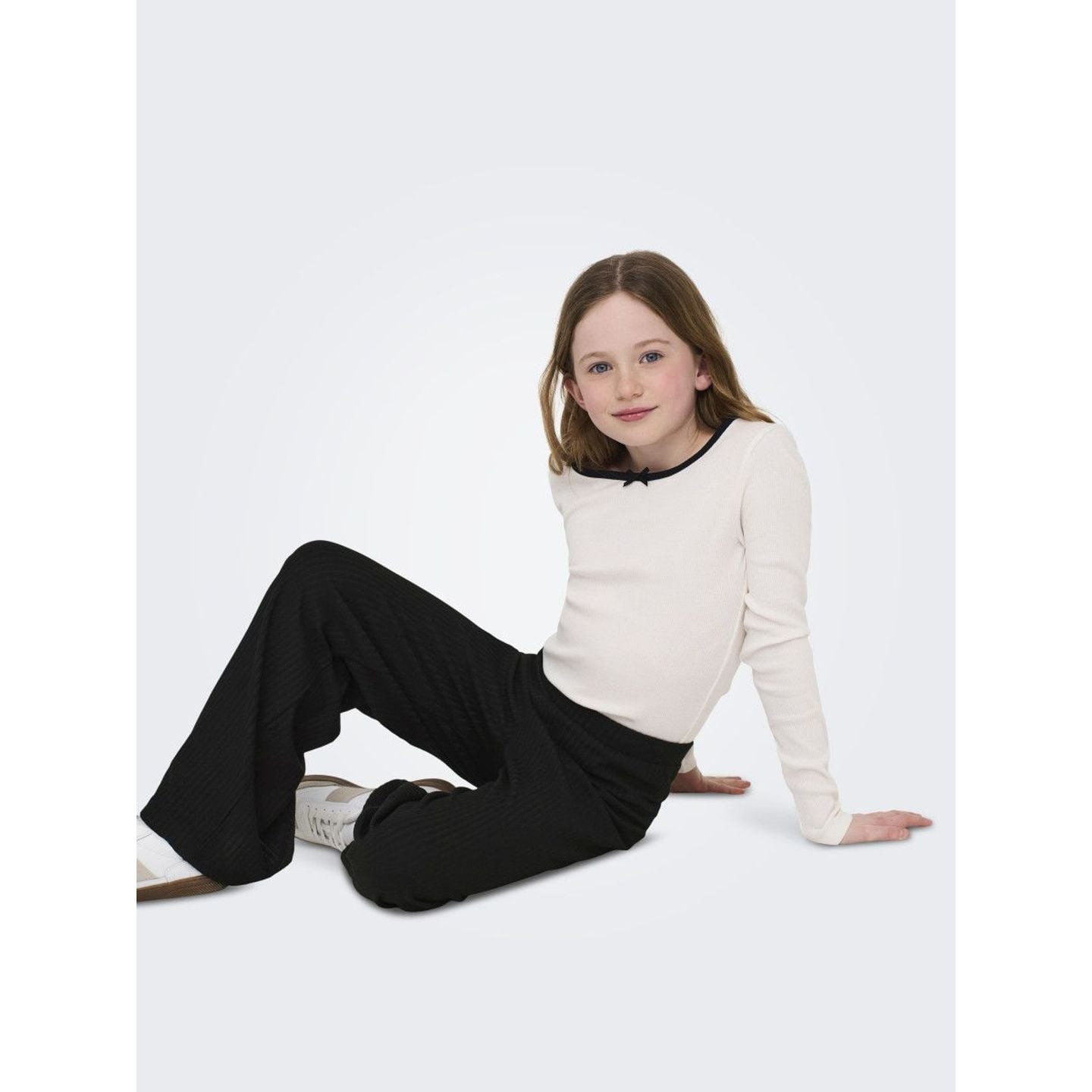 Fille assise au sol portant un pantalon large KOGNELLA PANT JRS NOOS noir, un haut blanc et des bask