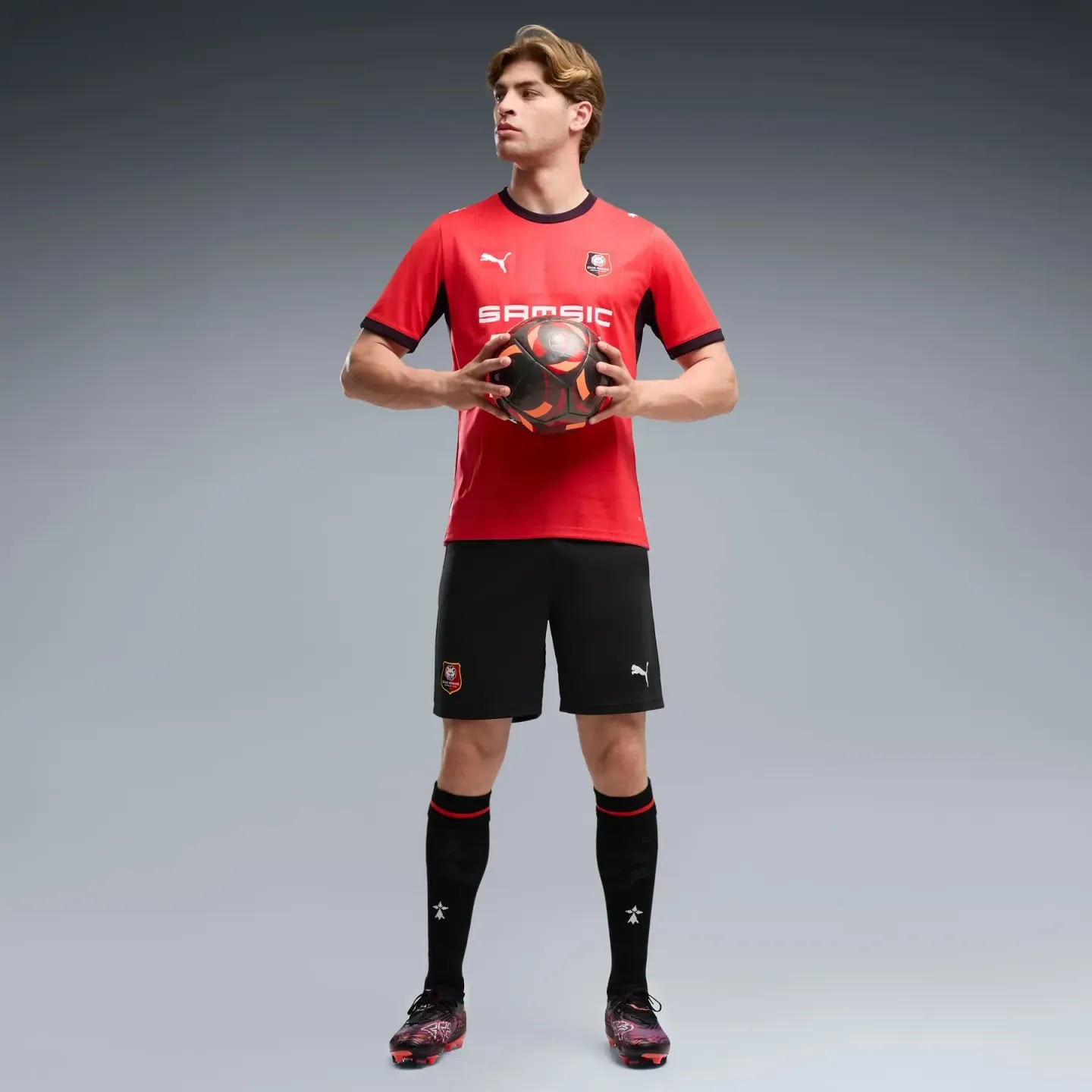 Footballeur portant le short réplique PUMA SRFC noir et rouge, tenant un ballon en studio